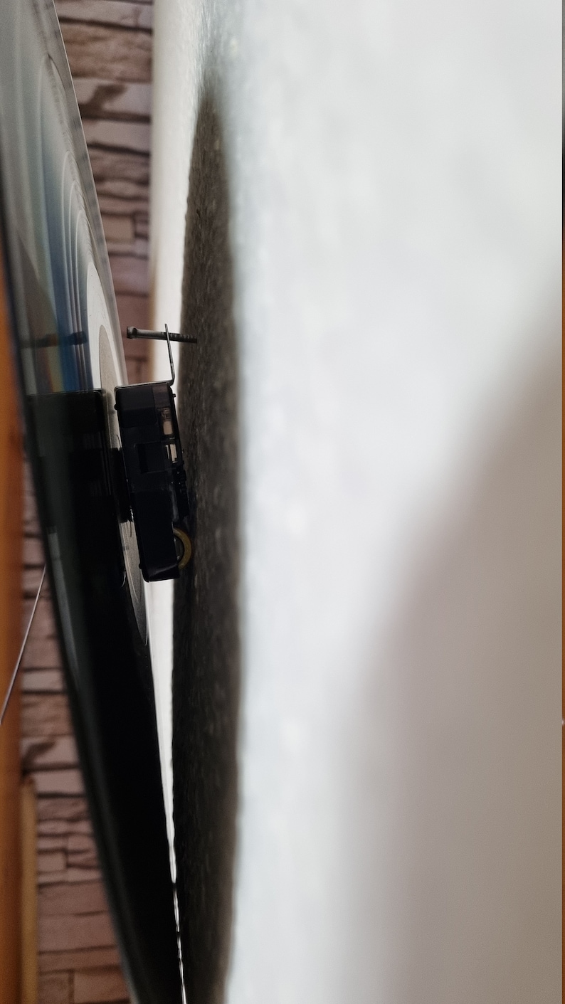 May include: Close-up of a black clock mechanism mounted on a wall. The clock mechanism has a silver hand and a black metal frame.