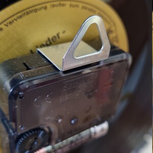 May include: Close-up of the back of a black clock with a silver hanging hook. The clock has a battery compartment and a gear visible. The clock is mounted on a black vinyl record.