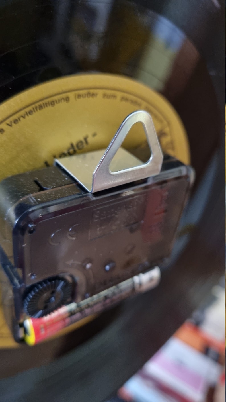 May include: Close-up of the back of a black clock with a silver hanging hook. The clock has a battery compartment and a gear visible. The clock is mounted on a black vinyl record.