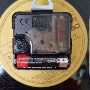 May include: Close-up of a black clock mechanism with a silver hanger. The clock face reads "Sang talo 61688" and "QUARTZ". The clock is powered by two AAA batteries.