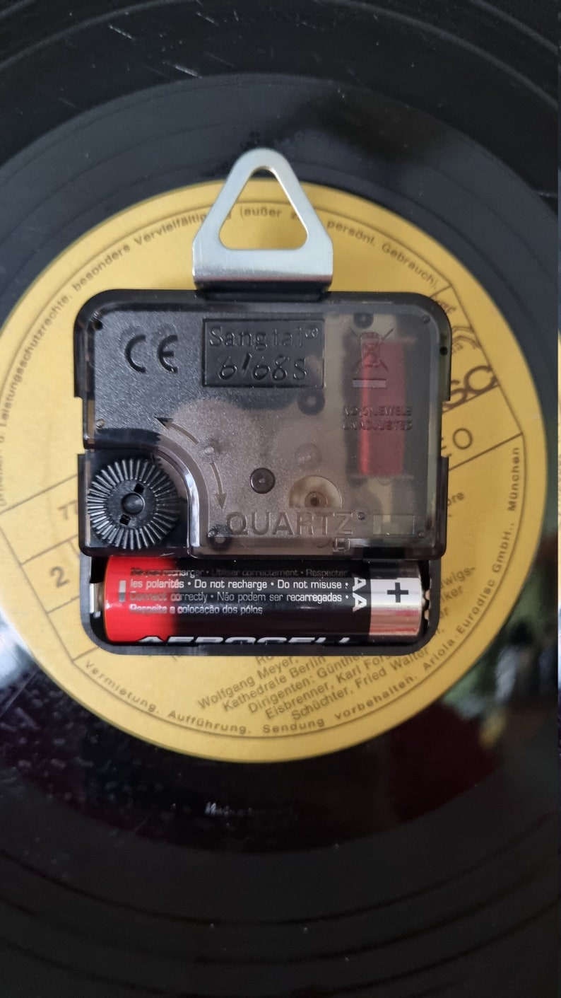 May include: Close-up of a black clock mechanism with a silver hanger. The clock face reads "Sang talo 61688" and "QUARTZ". The clock is powered by two AAA batteries.