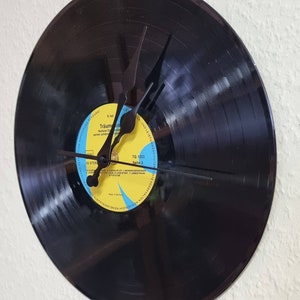 May include: A black vinyl record clock with black hands. The record label is yellow with blue text that reads "Traumreise" and "Seite 2".