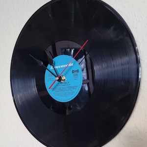 May include: A black vinyl record clock with a blue label. The label has the text "Accarola" and "JL. SILENZIO" on it. The clock hands are black and red.