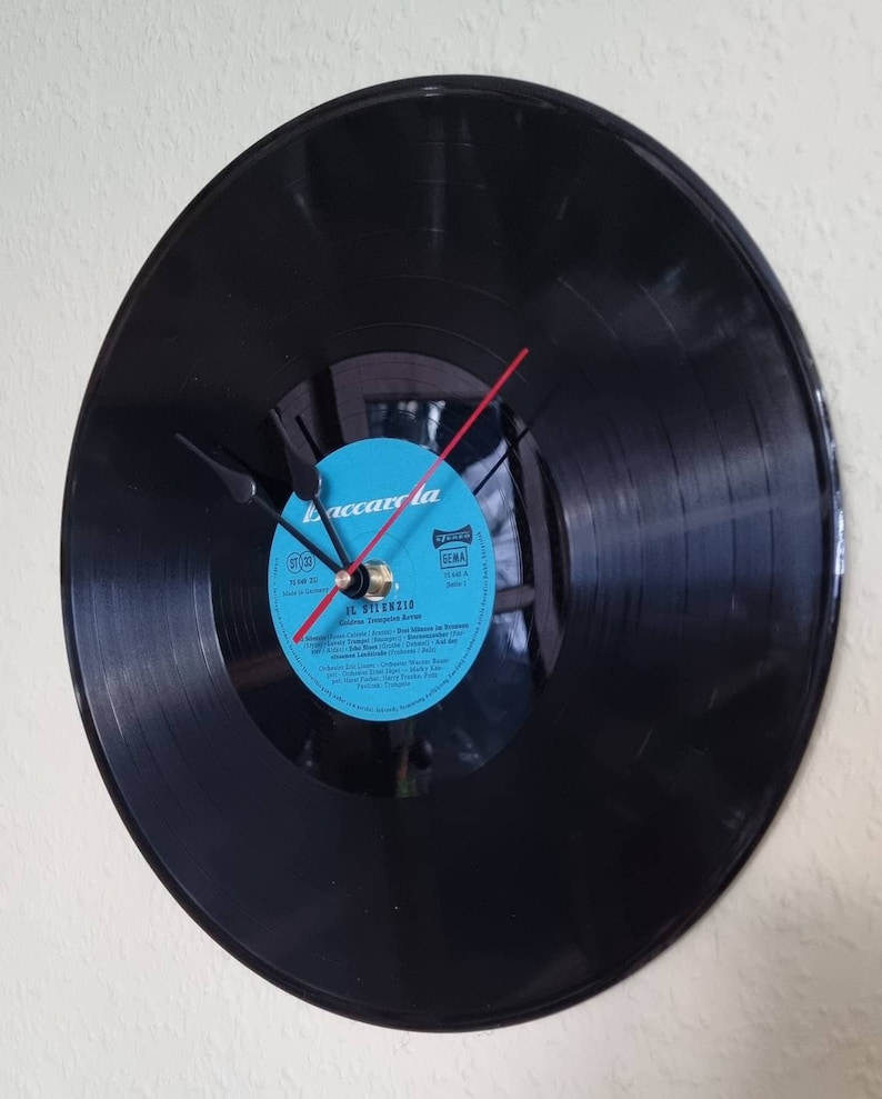 May include: A black vinyl record clock with a blue label. The label has the text "Accarola" and "JL. SILENZIO" on it. The clock hands are black and red.