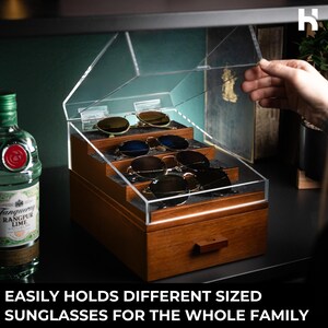 May include: A wooden sunglasses storage organizer with a clear acrylic hinged lid and a tiered interior featuring grey felt lining. The box includes a bottom drawer with a wooden handle and holds four pairs of sunglasses. Text reads: EASILY HOLDS DIFFERENT SIZED SUNGLASSES FOR THE WHOLE FAMILY.
