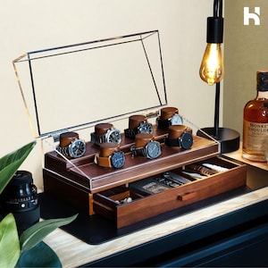 May include: A wooden watch box with a clear acrylic lid. The box has a drawer and multiple compartments for storing watches. The box is made of dark wood with a brown stain.