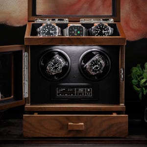 May include: A wooden watch display case with a glass top and door, showcasing several wristwatches. The case has two watch winders and a digital display. The watches have black and silver bands and faces. The case is made of dark wood.