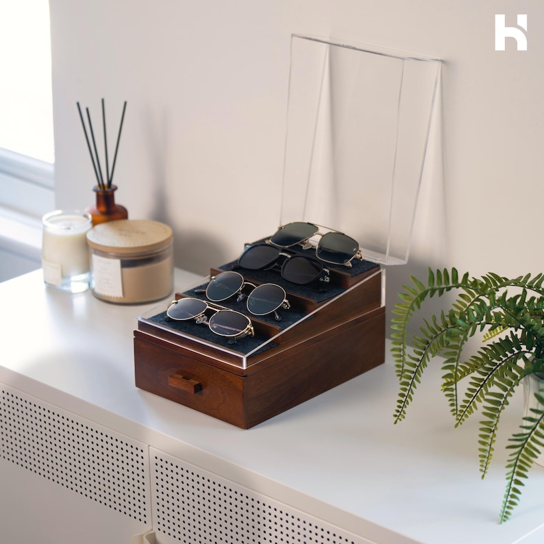 May include: A dark brown wooden storage box with a clear acrylic lid and a tiered interior display. The organizer features four slots lined with dark fabric, each holding a pair of sunglasses. The box includes a small drawer with a wooden handle.