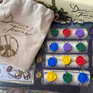 May include: A board game called "Darwin's Journey" featuring a canvas bag and game components. The bag is beige and has the game title printed on it. The game pieces include colourful wax seals and numbered cards.