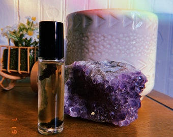 May include: A clear glass roller bottle with a black cap filled with a dark liquid. The bottle is sitting on a wooden surface next to a large purple amethyst crystal.