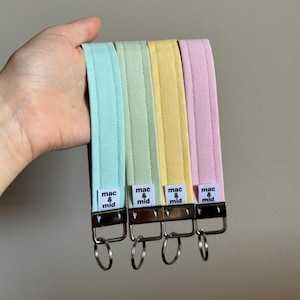 May include: Four pastel key fobs in blue, green, yellow, and pink. Each has a metal clasp with a key ring and a white label reading "mac & mid". The fobs are held in a hand against a neutral background.