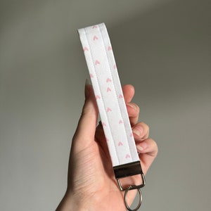 May include: White fabric wristlet with a pattern of small pink hearts. The wristlet has a silver-colored metal clasp and a silver key ring attached. The fabric is quilted with vertical stitching.