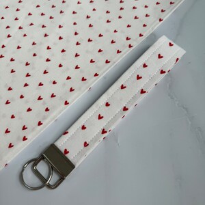 May include: A white fabric keychain wristlet with a pattern of small red hearts. The wristlet has a silver-colored metal clasp and a key ring. A matching fabric piece is visible in the background, also covered in red hearts.