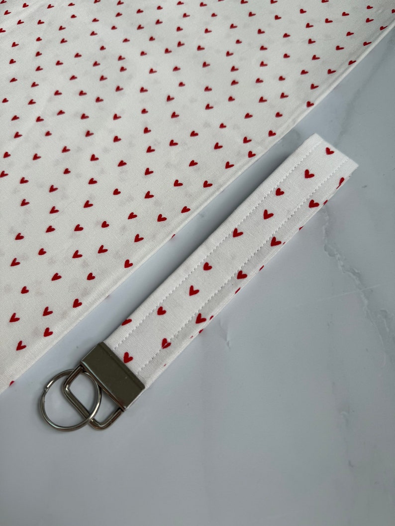 May include: A white fabric keychain wristlet with a pattern of small red hearts. The wristlet has a silver-colored metal clasp and a key ring. A matching fabric piece is visible in the background, also covered in red hearts.