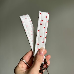 May include: White fabric wrist lanyard with a repeating pattern of small red hearts. The lanyard has a metal clasp with two key rings. The fabric is quilted with a pink heart pattern on the back.