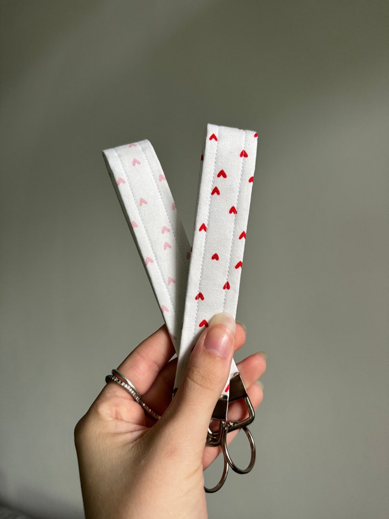 May include: White fabric wrist lanyard with a repeating pattern of small red hearts. The lanyard has a metal clasp with two key rings. The fabric is quilted with a pink heart pattern on the back.