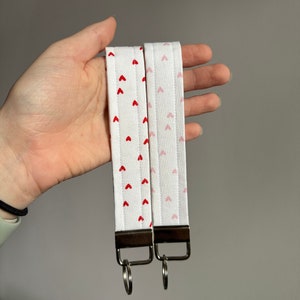May include: Two white fabric wrist keychains with red and pink heart patterns. Each keychain has a silver-colored metal loop and a rectangular metal piece. The keychains are held in a hand.