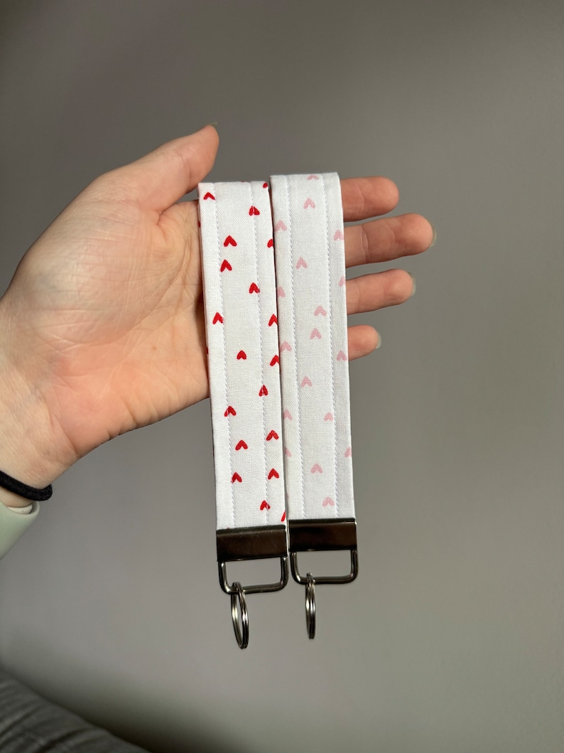 May include: Two white fabric wrist keychains with red and pink heart patterns. Each keychain has a silver-colored metal loop and a rectangular metal piece. The keychains are held in a hand.