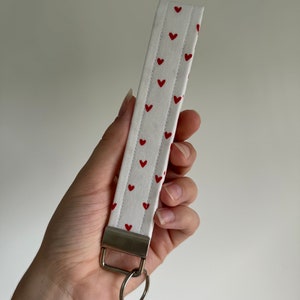 May include: White fabric wristlet with a pattern of small red hearts. The wristlet has a metal clasp and a silver key ring attached. The fabric is quilted with vertical stitching.