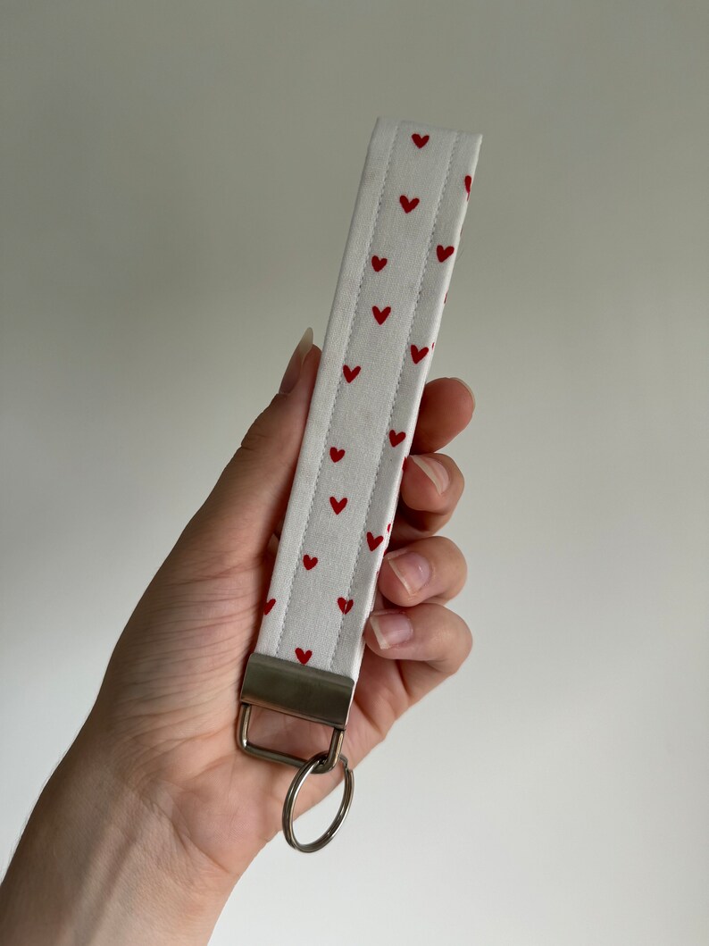 May include: White fabric wristlet with a pattern of small red hearts. The wristlet has a metal clasp and a silver key ring attached. The fabric is quilted with vertical stitching.