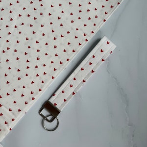 May include: White fabric with a repeating pattern of small red hearts. A matching key fob with a metal ring is also visible. The key fob is white with red heart accents and a metal ring.