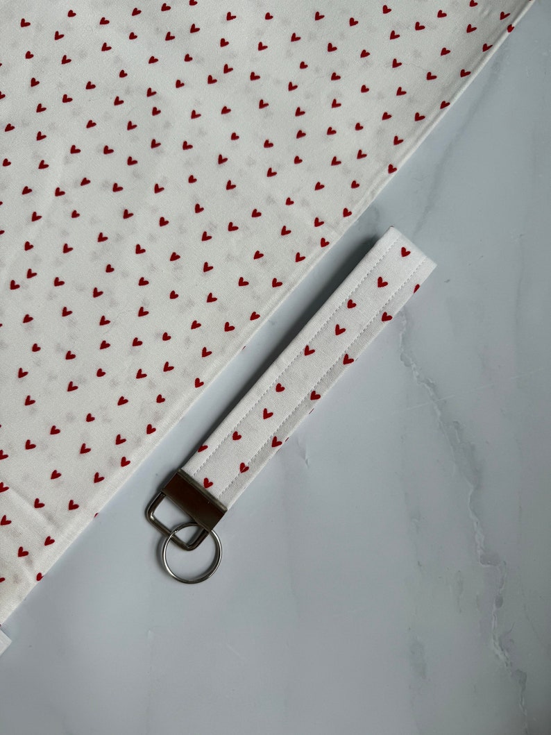 May include: White fabric with a repeating pattern of small red hearts. A matching key fob with a metal ring is also visible. The key fob is white with red heart accents and a metal ring.