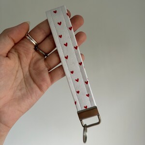 May include: White fabric wristlet key fob with a pattern of small red hearts. The key fob has a metal clasp and a silver key ring attached. The wristlet is held in a hand against a white background.