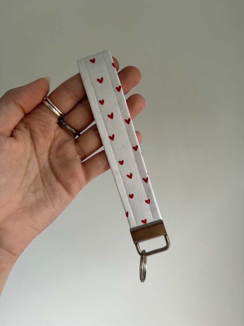 May include: White fabric wristlet key fob with a pattern of small red hearts. The key fob has a metal clasp and a silver key ring attached. The wristlet is held in a hand against a white background.