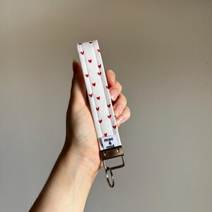 May include: White fabric wristlet with a pattern of small red hearts. The wristlet has a silver-colored metal clasp and a small tag that reads "mac & mid". A silver key ring is attached to the clasp.