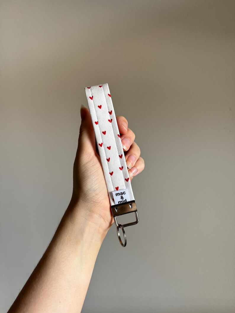 May include: White fabric wristlet with a pattern of small red hearts. The wristlet has a silver-colored metal clasp and a small tag that reads "mac & mid". A silver key ring is attached to the clasp.