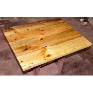 Peut inclure: Une table carrée en bois avec une finition naturelle. Le plateau est en bois clair avec des veines et des nœuds visibles. La table est soutenue par un piètement peint en rouge. La table mesure environ 76 cm de côté.