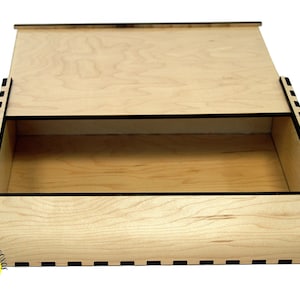 May include: A wooden box with a sliding lid, made from light-coloured wood. The box is rectangular with a visible interior and black accents. The lid is partially open, revealing the inside. The box is likely for storage, possibly for crafts or small items.