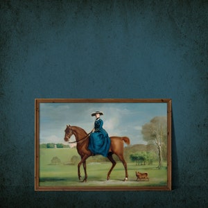 May include: A framed oil painting depicting a woman in a blue dress riding a brown horse. The woman is wearing a wide-brimmed hat and holding the reins. The horse is walking through a grassy field with a small dog running behind it.