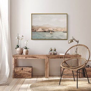 Op de afbeelding: Een licht houten bureau met een gouden lijst met een schilderij van een zeegezicht erboven. Een rieten stoel met een gouden metalen frame staat voor het bureau. Er staan twee planten in potten op het bureau.