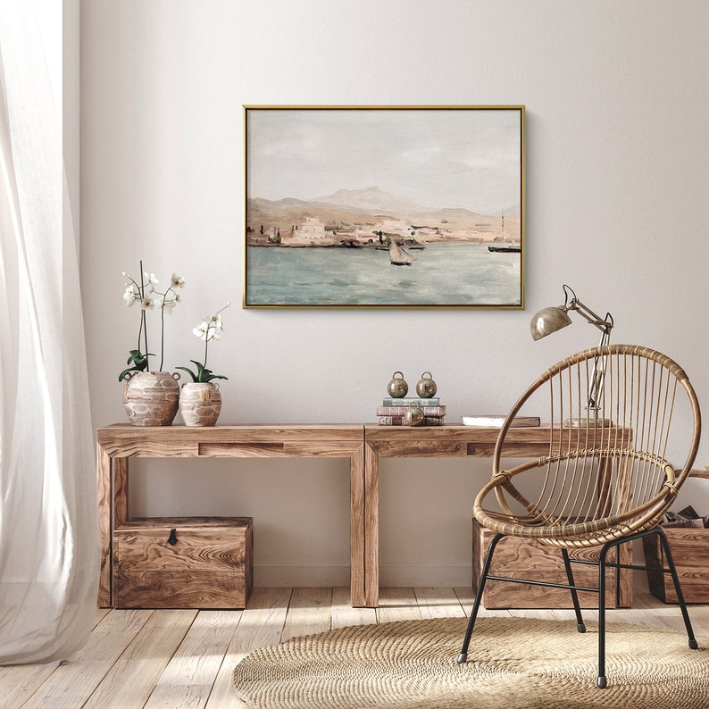 Op de afbeelding: Een licht houten bureau met een gouden lijst met een schilderij van een zeegezicht erboven. Een rieten stoel met een gouden metalen frame staat voor het bureau. Er staan twee planten in potten op het bureau.