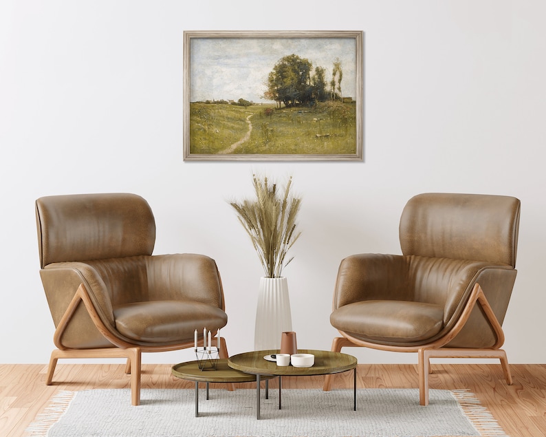 May include: Two brown leather armchairs with wooden frames, facing each other in a living room setting. A framed landscape painting hangs on the wall behind the chairs. A vase with dried wheat stalks sits on a coffee table between the chairs.