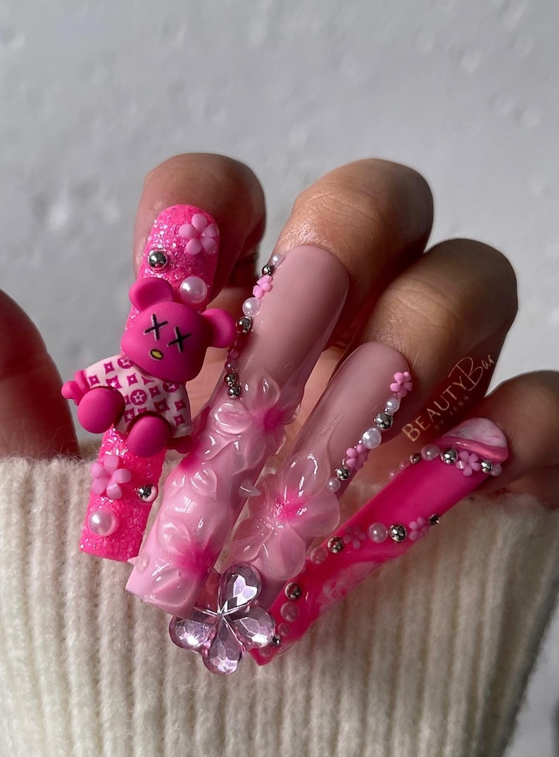 Puede incluir: U&ntilde;as acr&iacute;licas largas de color rosa y blanco con flores 3D, pedrer&iacute;a y un dije de oso de peluche rosa. Las u&ntilde;as est&aacute;n decoradas con perlas y cuentas plateadas. El texto "BEAUTYBAR" es visible en una de las u&ntilde;as.