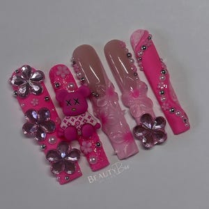 Puede incluir: U&ntilde;as postizas rosas y blancas con un dije de oso de peluche rosa, flores rosas y pedrer&iacute;a. Las u&ntilde;as est&aacute;n decoradas con un dise&ntilde;o floral rosa y blanco y un dise&ntilde;o de remolino rosa y blanco. Las u&ntilde;as est&aacute;n etiquetadas como "BEAUTYBar."