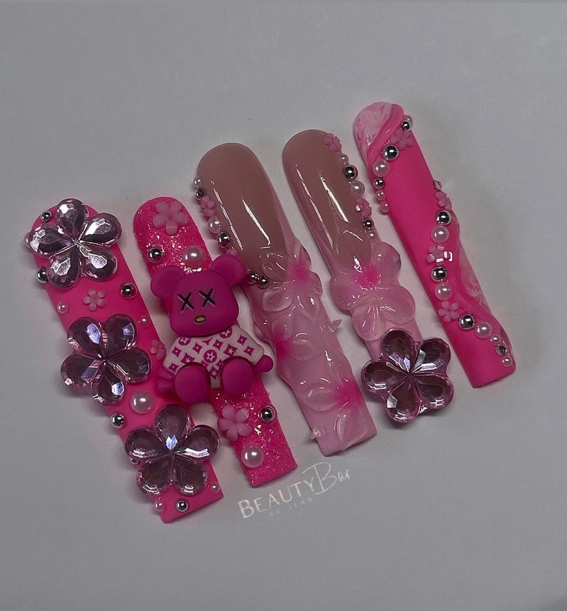 Puede incluir: U&ntilde;as postizas rosas y blancas con un dije de oso de peluche rosa, flores rosas y pedrer&iacute;a. Las u&ntilde;as est&aacute;n decoradas con un dise&ntilde;o floral rosa y blanco y un dise&ntilde;o de remolino rosa y blanco. Las u&ntilde;as est&aacute;n etiquetadas como "BEAUTYBar."