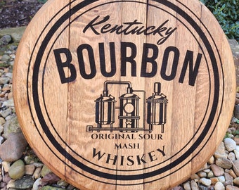 May include: Round wooden sign with the words "Kentucky Bourbon" in a black, bold font. The sign also features an illustration of a distillery and the words "Original Sour Mash Whiskey". The sign is made of wooden barrel staves.