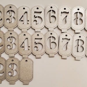 May include: Set of twenty silver-coloured wooden tags with numbers 1 through 10, each number appearing twice. The tags are shaped like a rectangle with rounded corners and have a hole at the top for hanging.