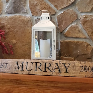 May include: A wooden sign with the text "EST. MURRAY 2000" engraved on it. The sign is mounted on a rustic wooden surface with metal brackets on each end. A white lantern is sitting on the surface behind the sign.