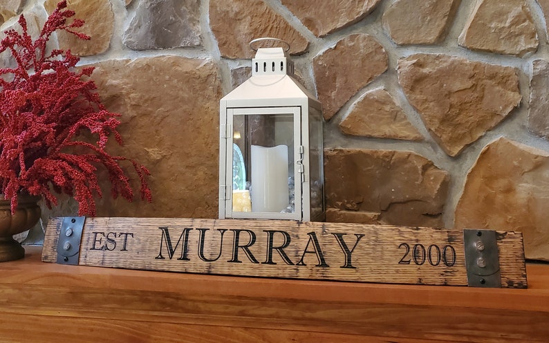 May include: A wooden sign with the text "EST. MURRAY 2000" engraved on it. The sign is mounted on a rustic wooden surface with metal brackets on each end. A white lantern is sitting on the surface behind the sign.