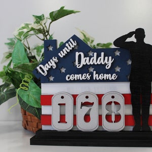 May include: A wooden house-shaped countdown plaque with an American flag design. It says "Days until Daddy comes home" and features a silhouette of a soldier saluting. The numbers on the plaque read "178".