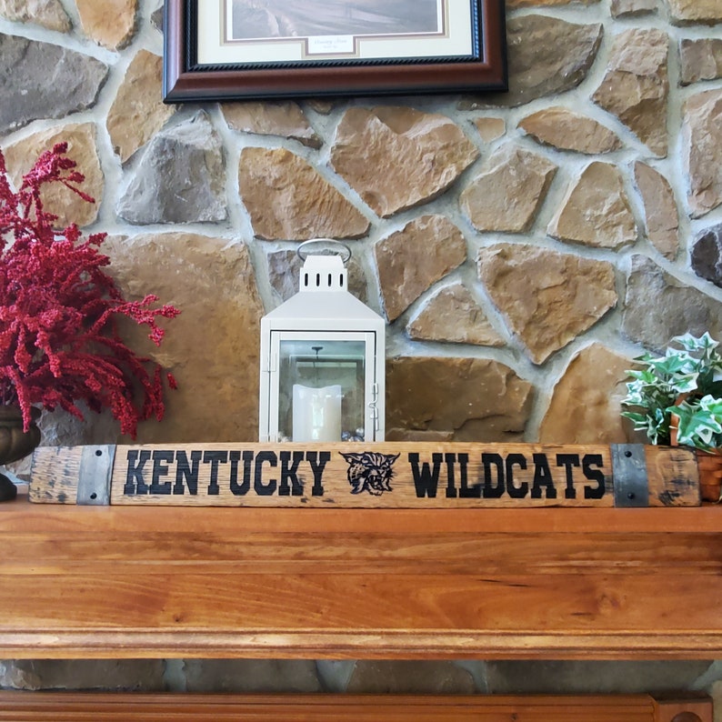 May include: A wooden sign with the words "Kentucky Wildcats" in black lettering. The sign is decorated with a black and white image of a wildcat. The sign is sitting on a wooden mantel above a stone fireplace.