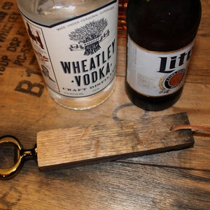 May include: A wooden bottle opener with a gold metal opener and a leather strap. The bottle opener is sitting on a wooden surface with two bottles of alcohol in the background. The bottles are labelled "Wheatley Vodka" and "Lite".