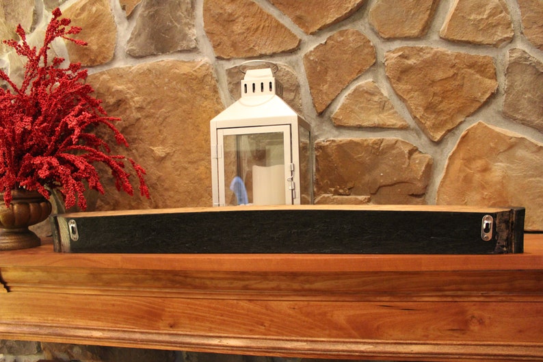 May include: A black wooden shelf made from a reclaimed wine barrel with metal brackets for hanging on a wall. The shelf is mounted on a wooden mantel above a stone fireplace.