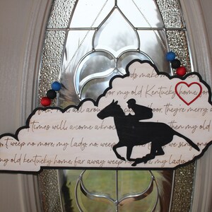 May include: A wooden Kentucky state-shaped sign with a black silhouette of a horse and rider. The sign has a red heart with the word "home" written inside. The sign has the lyrics to the song "My Old Kentucky Home" written on it.