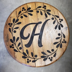 May include: A round wooden sign with a black painted wreath and the letter "H" in the centre. The wreath is made of stylised leaves and berries.
