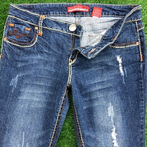 Könnte beinhalten: Blaue Jeans aus Denim mit Destroyed-Details und hellbraunen Nähten. Die Jeans haben einen Reißverschluss und einen Knopfverschluss. Der Bund hat ein rotes Etikett mit dem Text "Java Di Jean 13".